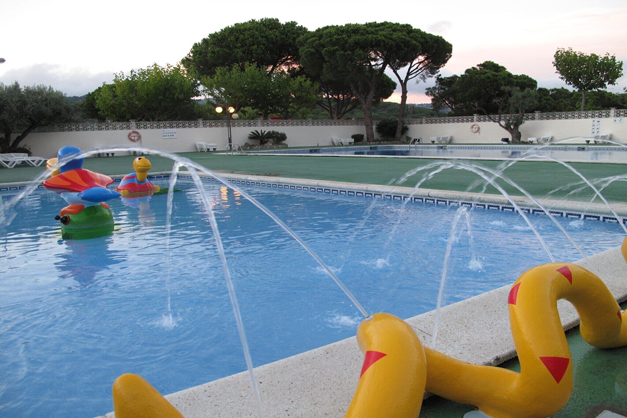 En swimmingpool med vattenfontäner, uppblåsbara leksaker och utsikt över träd och en vägg i bakgrunden.