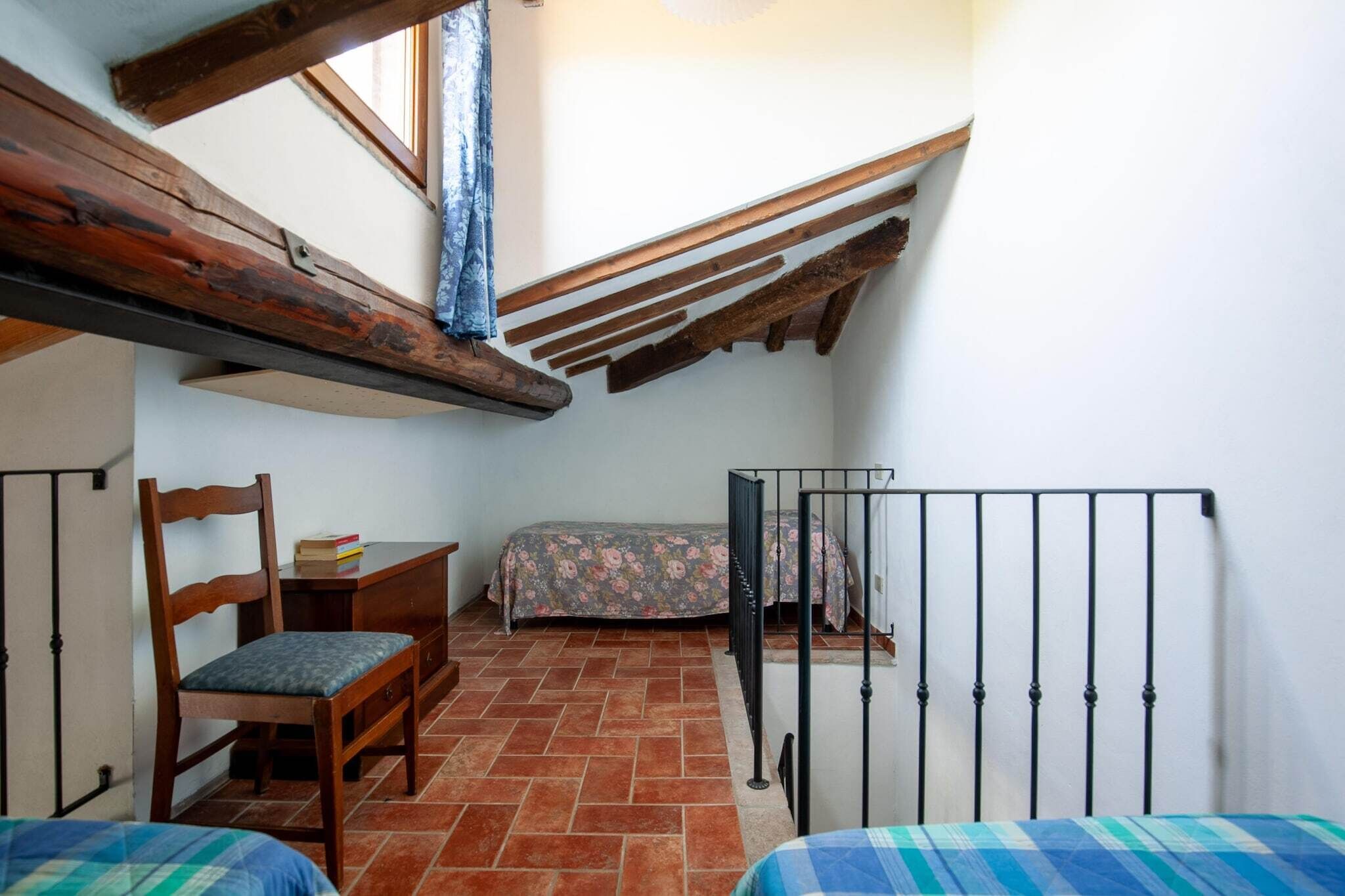 Una piccola stanza rustica con soffitto spiovente, una sedia di legno, un letto con struttura metallica e una scrivania con una sedia.