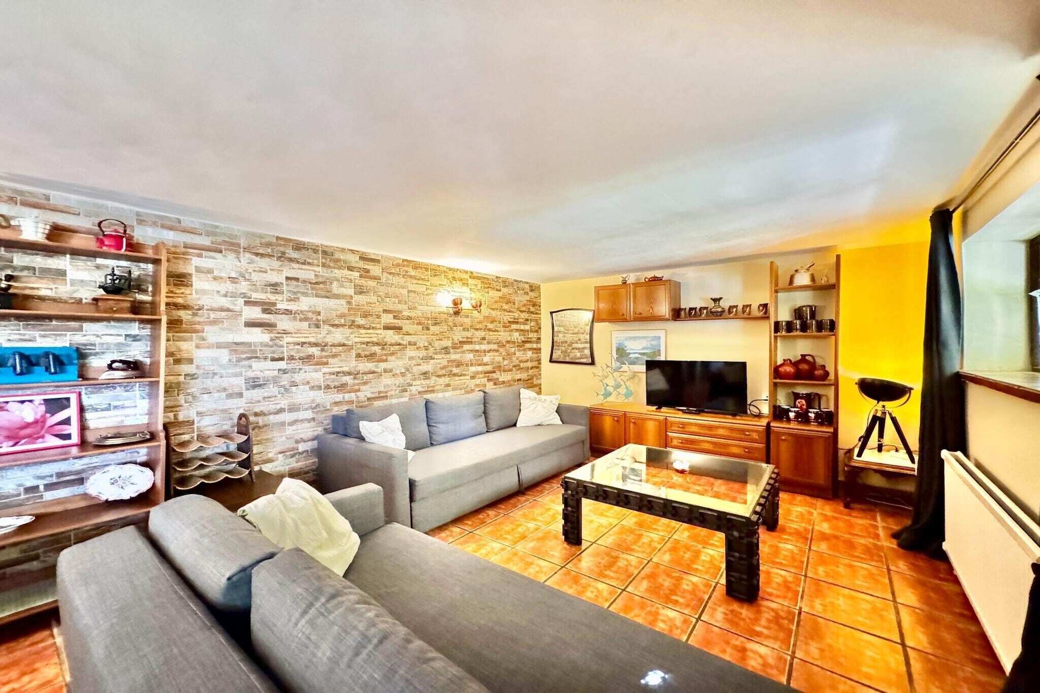A living room with a grey sofa, a coffee table, and a TV. There is a kitchen area with cabinets and a stove.