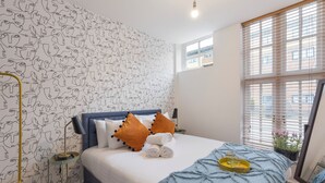 A neatly made bed with a blue blanket, white pillows, and orange accent pillows. A bedside table holds a lamp and a tray with candles. The room features a patterned wallpaper and a window with blinds. - Amazingly Beautiful 3 BR Flat For Large Groups (London)