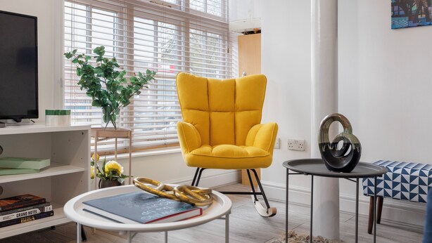 A modern living room with a yellow armchair, a round coffee table with books, a small side table with a vase, and a painting on the wall. - Amazingly Beautiful 3 BR Flat For Large Groups (London)