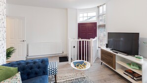 A modern living room with a blue sofa, a small round table with books, a television on a white stand, and a red door. - Amazingly Beautiful 3 BR Flat For Large Groups (London)