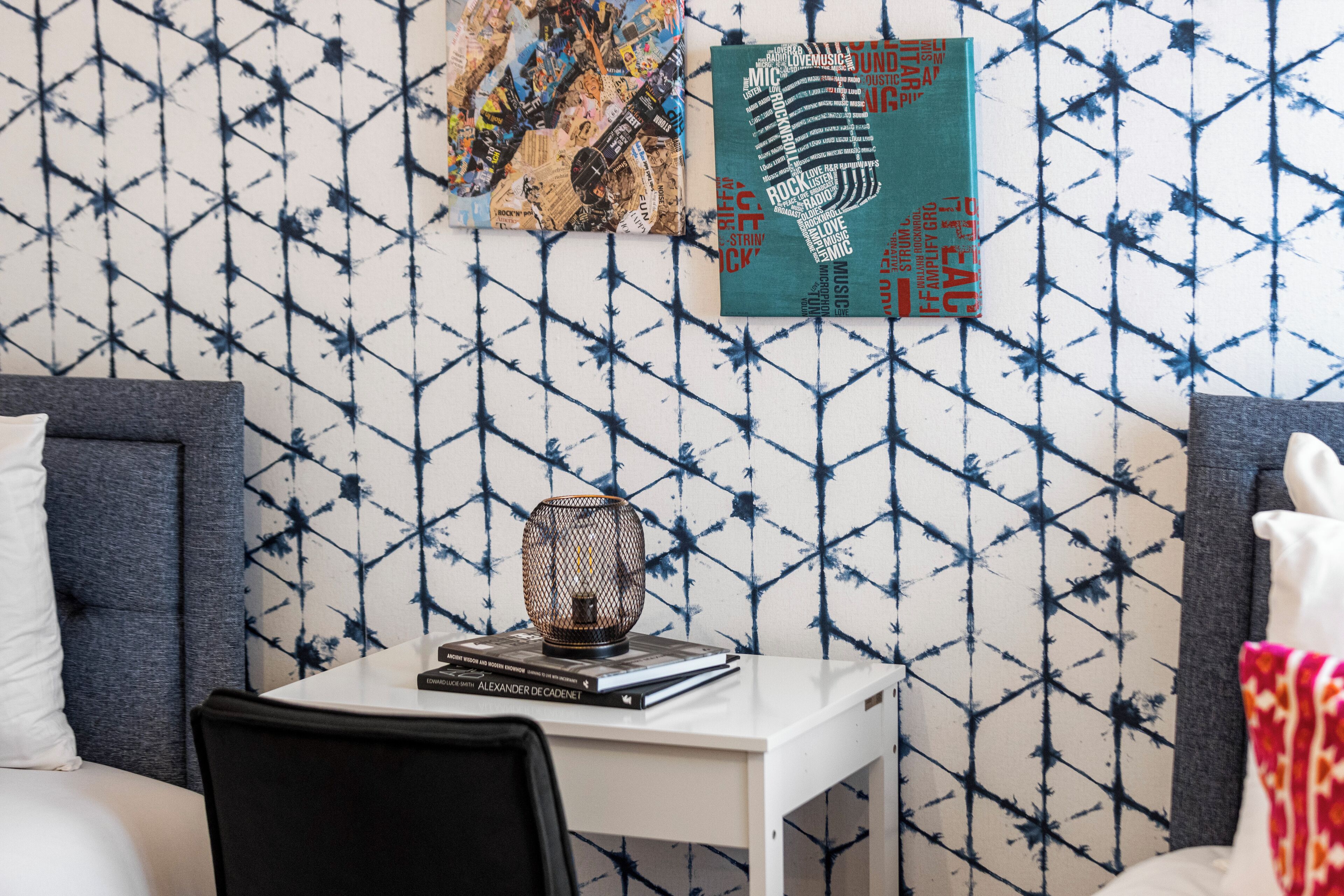 Une chambre d’hôtel avec un papier peint à motifs géométriques, une table de chevet blanche sur laquelle se trouvent un livre et une lampe décorative, et un lit avec une tête de lit grise et des oreillers blancs.