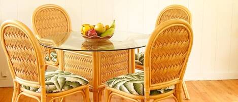 Un comedor con una mesa redonda de vidrio rodeada por cuatro sillas de mimbre con asientos acolchados. Hay un frutero sobre la mesa.