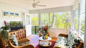 Amplia sala de estar con televisor, mobiliario de mimbre y ventanas grandes con vistas al verdor.