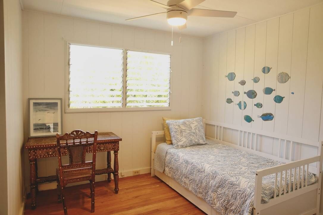 A bedroom with a bed, a desk, and a chair. There is a window with blinds and a ceiling fan.