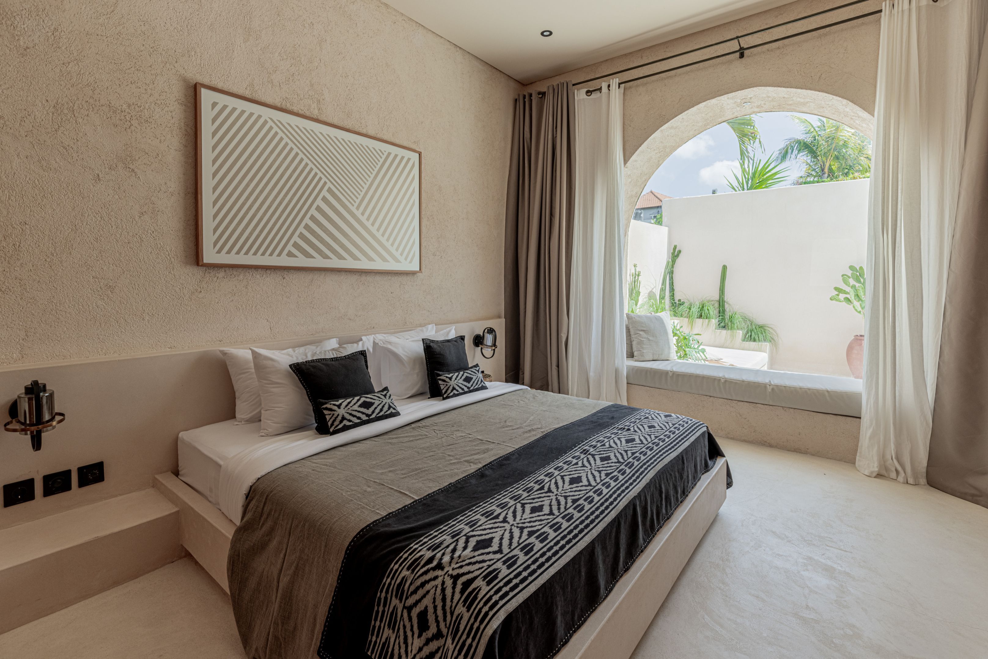 A modern bedroom with a large bed, a geometric wall art piece, and a view of a courtyard through a window with curtains.