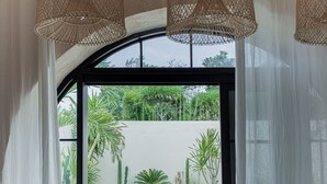 A modern interior with a view of an outdoor pool area, featuring a large window, wicker ceiling lamps, and a rustic wooden table. - Khaleela Villas (Canggu)