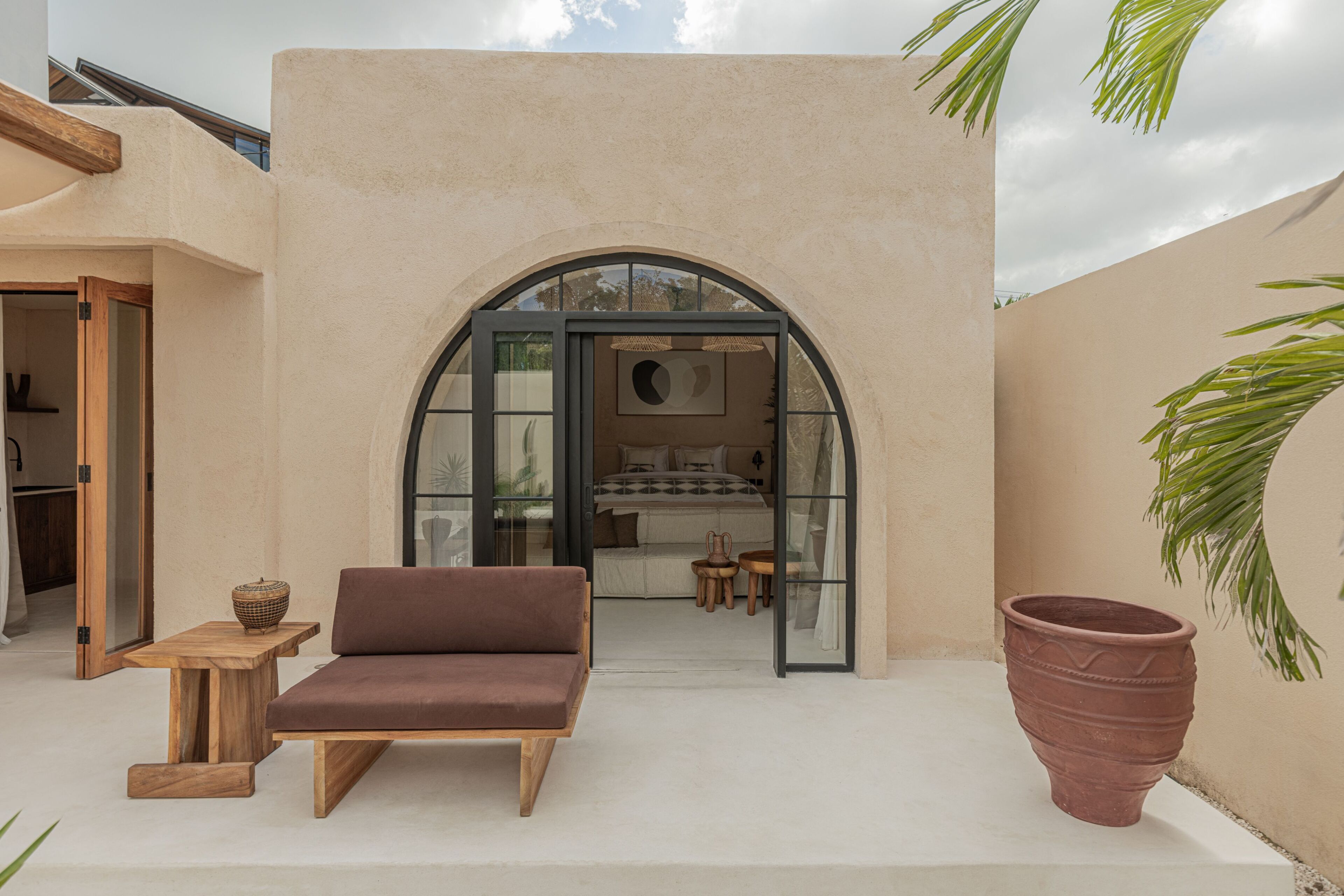 A cozy outdoor seating area with a sofa and a table, a large terracotta pot, and a view into a bedroom through an arched doorway.