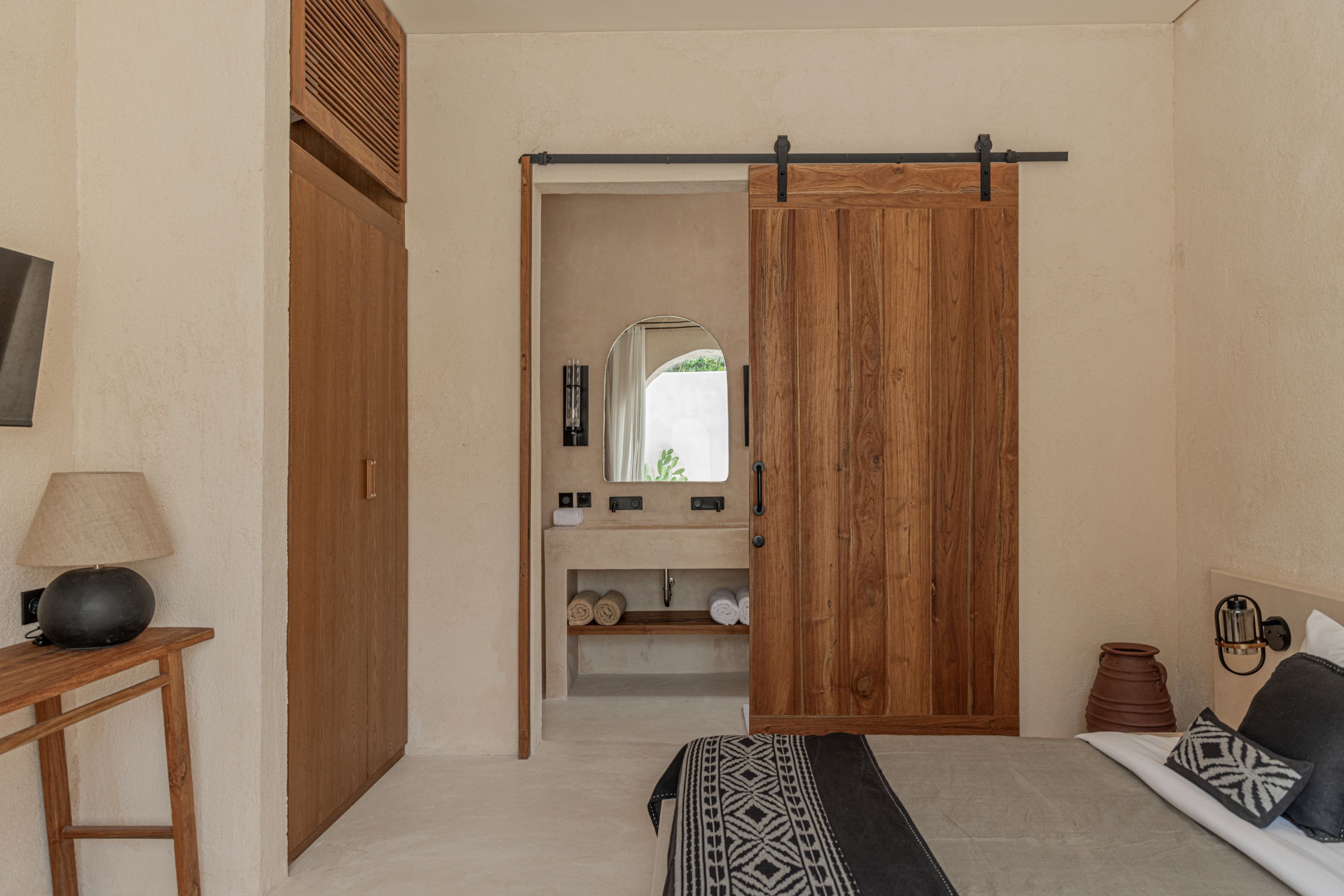 A bedroom with a wooden wardrobe, a bedside table with a lamp, a bed with patterned bedding, and a view through an open door to a bathroom with a mirror and shelves.