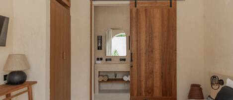 A bedroom with a wooden wardrobe, a bedside table with a lamp, a bed with patterned bedding, and a view through an open door to a bathroom with a mirror and shelves.