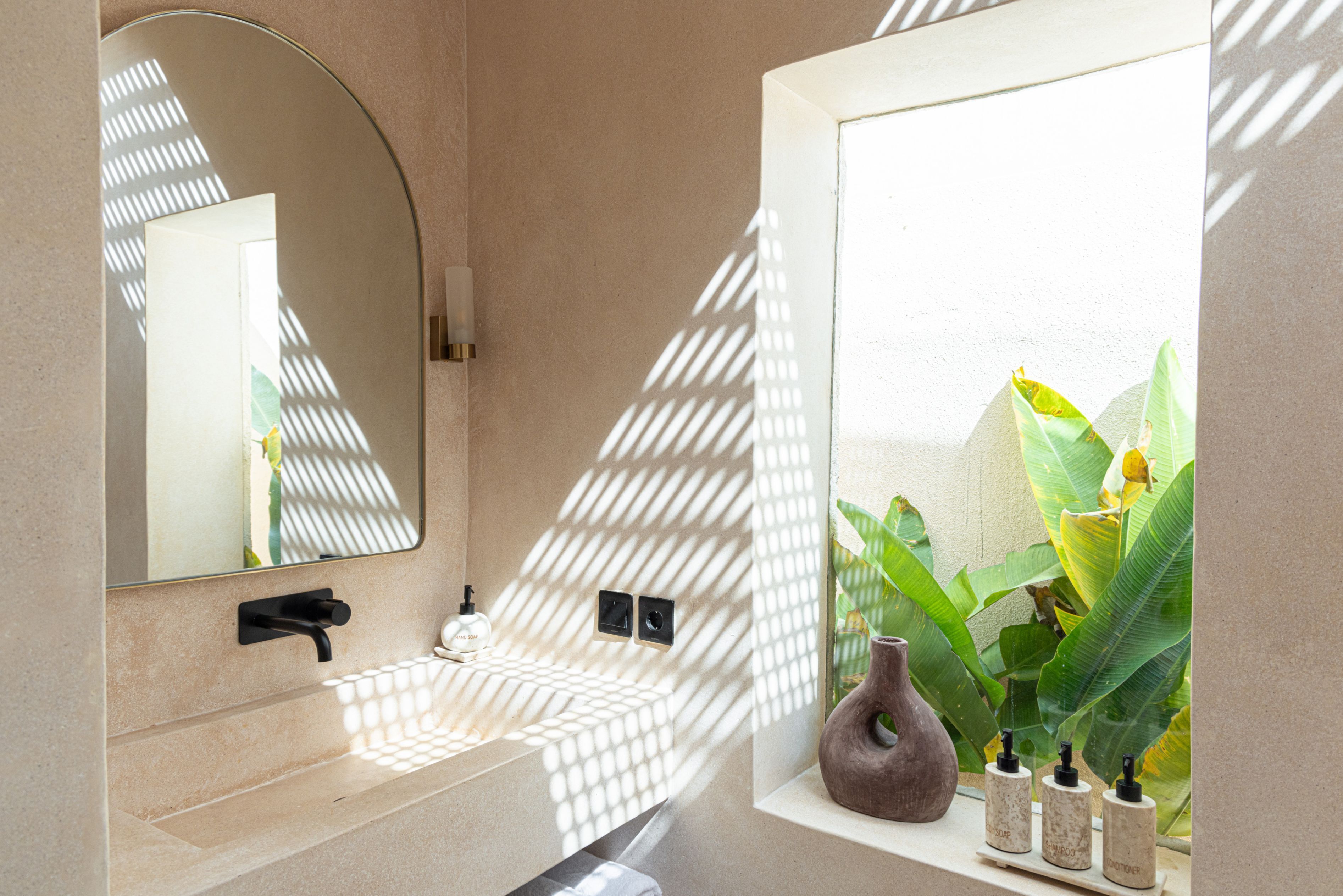 A bathroom with a beige countertop, a black faucet, a mirror, and a window with plants.