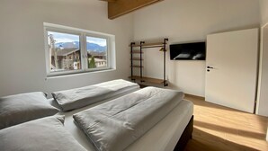 A bedroom with two beds, a window with a view of houses, a TV mounted on the wall, and a wardrobe.