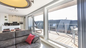 A modern living room with a grey sofa and a red pillow, a dining area with a table and chairs, and a balcony with a wooden deck and a view of the cityscape.