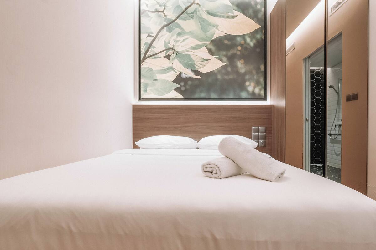 A neatly made bed with white linens and a rolled towel, a wooden headboard, and a window with a plant view.
