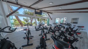 Uma academia com várias bicicletas ergométricas dispostas em fileiras, grandes janelas com vista para árvores e um corpo d'água, e piso de madeira.