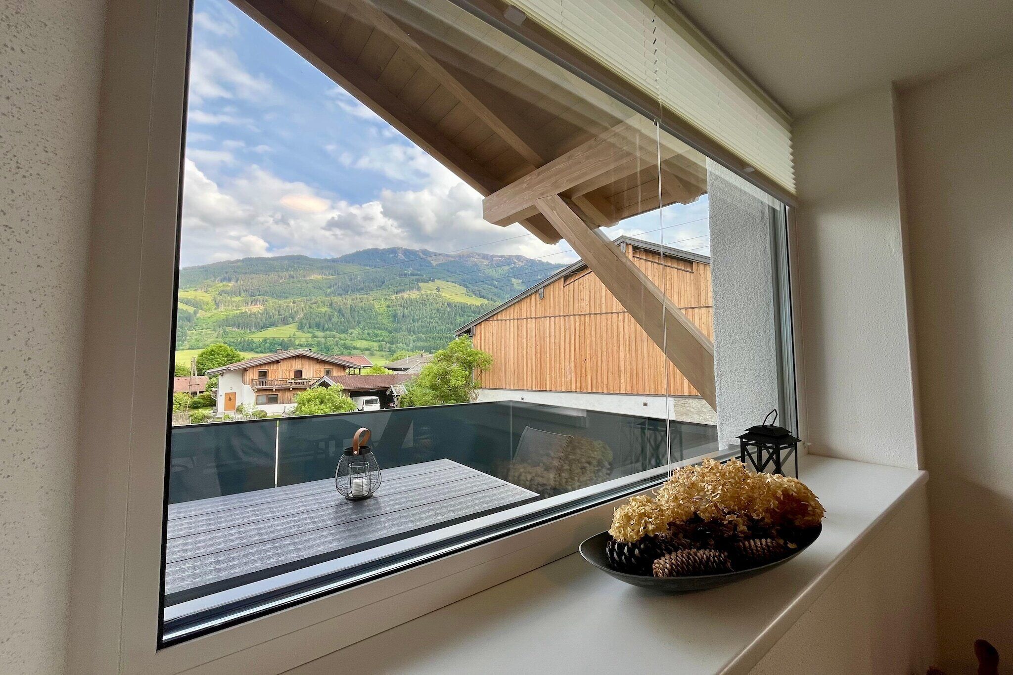Un balcón con vistas a un paisaje montañoso, baranda de madera, una mesita con un farol y flores.