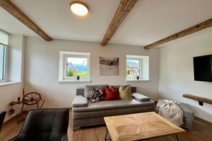 A modern living room with a sofa, a wooden coffee table, and a television. There are windows with views of greenery and a painting on the wall.