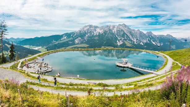 Un lago circular con un muelle, rodeado de montañas y un cielo despejado.