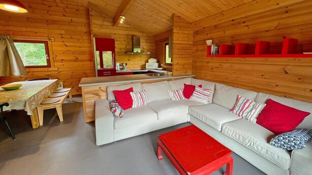 Un acogedor interior de cabaña con detalles de madera, un sofá blanco con cojines rojos, una mesa de centro roja y una zona de cocina al fondo.