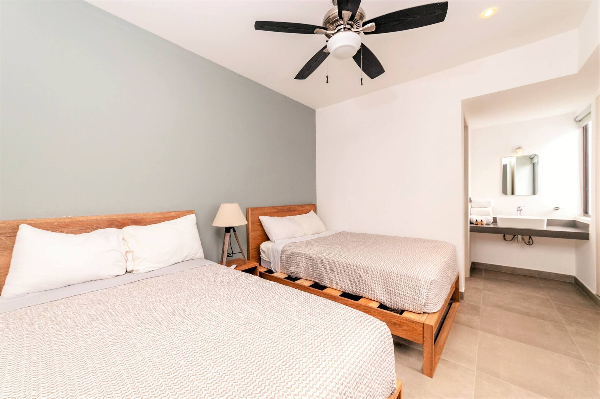 Une chambre avec deux lits, un ventilateur de plafond, une lampe de chevet et une salle de bain avec un lavabo et un miroir.