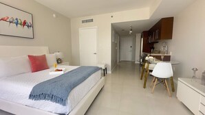 A modern bedroom with a bed, bedside table, and a painting. There is a kitchenette with a sink and cabinets.
