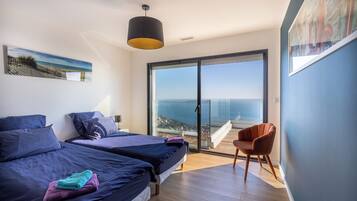 A modern bedroom with two beds, a large sliding glass door to a balcony, and a view of the sea.