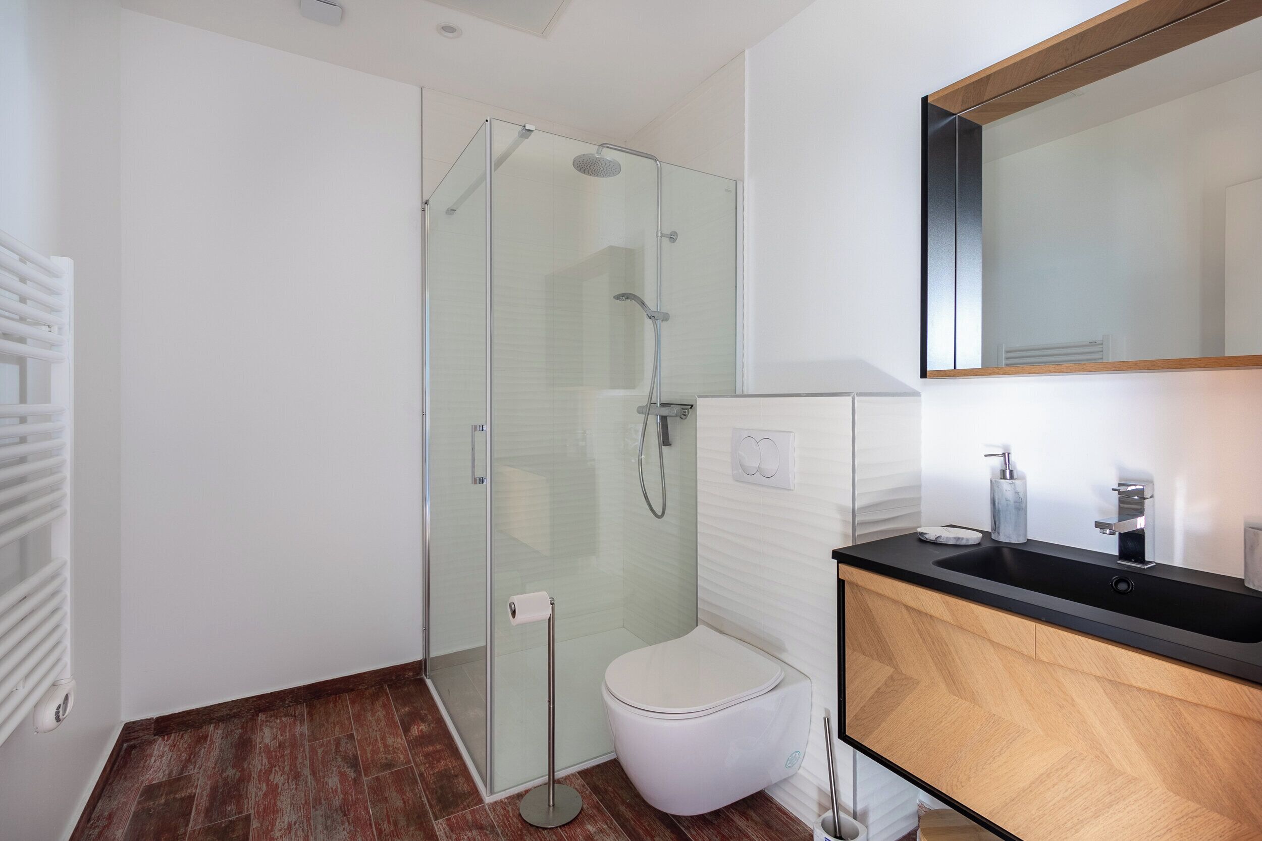 A modern bathroom with a glass-enclosed shower, a white toilet, and a wooden vanity with a black countertop.