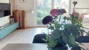 A modern living room with a wooden ceiling, a large window, and a dining area with a table and chairs. - 6P wooden house + garden on the Elbe dike | modern, close to nature, family & dog-friendly (Artlenburg)