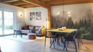 A spacious room with a wooden floor, a dining table with chairs, a sofa, a coffee table, and a mural of a forest on the wall. - 6P wooden house + garden on the Elbe dike | modern, close to nature, family & dog-friendly (Artlenburg)