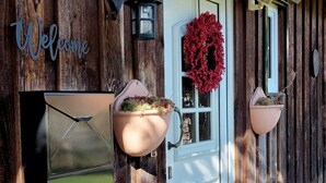 A rustic wooden building with a white door, a red wreath, and two hanging planters. - 6P wooden house + garden on the Elbe dike | modern, close to nature, family & dog-friendly (Artlenburg)