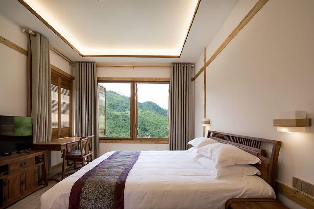 Ein Hotelzimmer mit Bett, Schreibtisch mit Stuhl, Fernseher und großen Fenstern mit Blick auf Grünflächen.