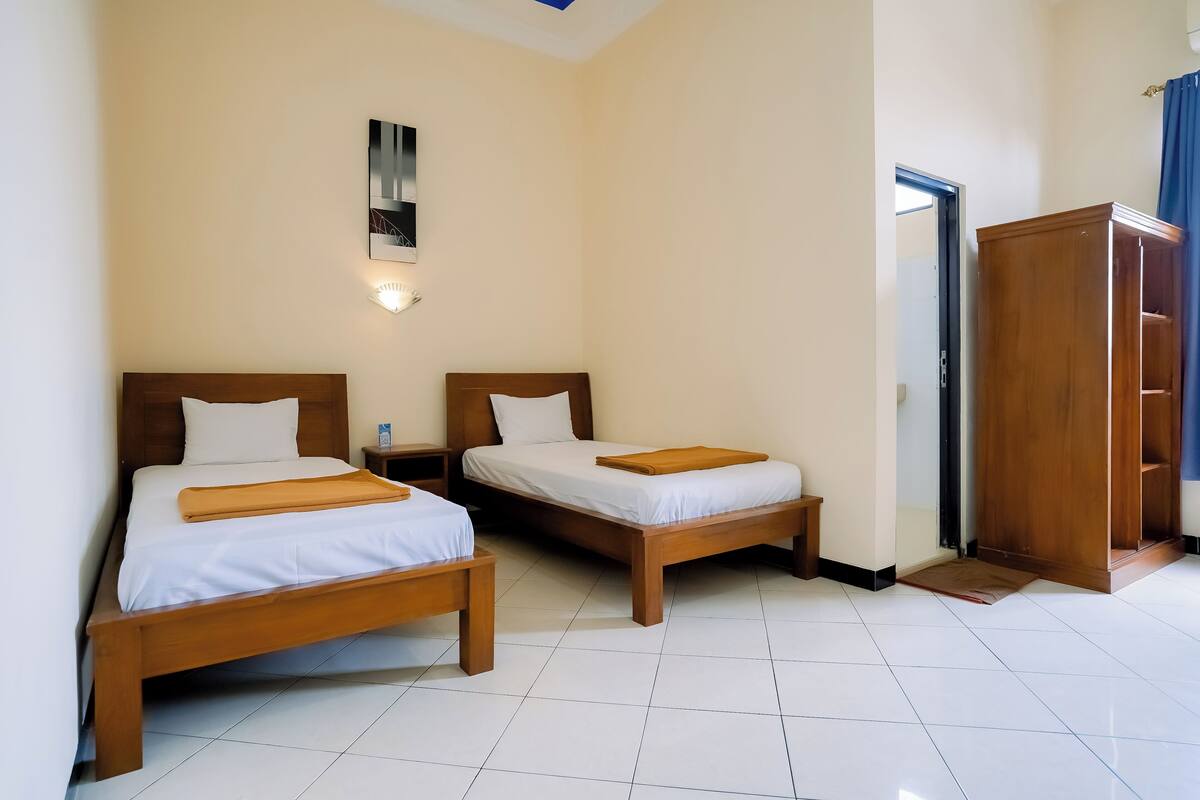 Quarto de hotel com duas camas de madeira, um guarda-roupa e uma mesa de cabeceira com abajur.