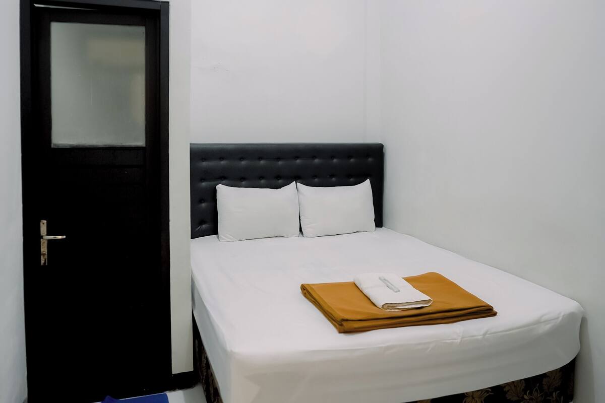 Quarto pequeno e bem arrumado, com uma cama, uma porta preta e um tapete azul no chão.