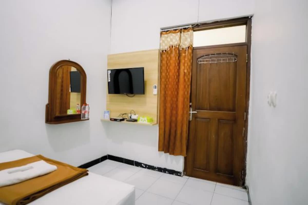 A hotel room with a bed, a wooden door, a mirror, a television, and a small shelf with toiletries.