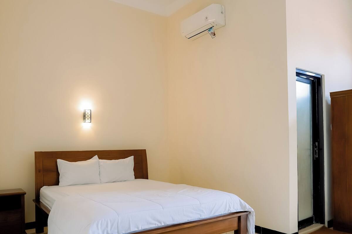 Quarto com cama de madeira, mesa de cabeceira, ar condicionado fixado na parede e uma porta.