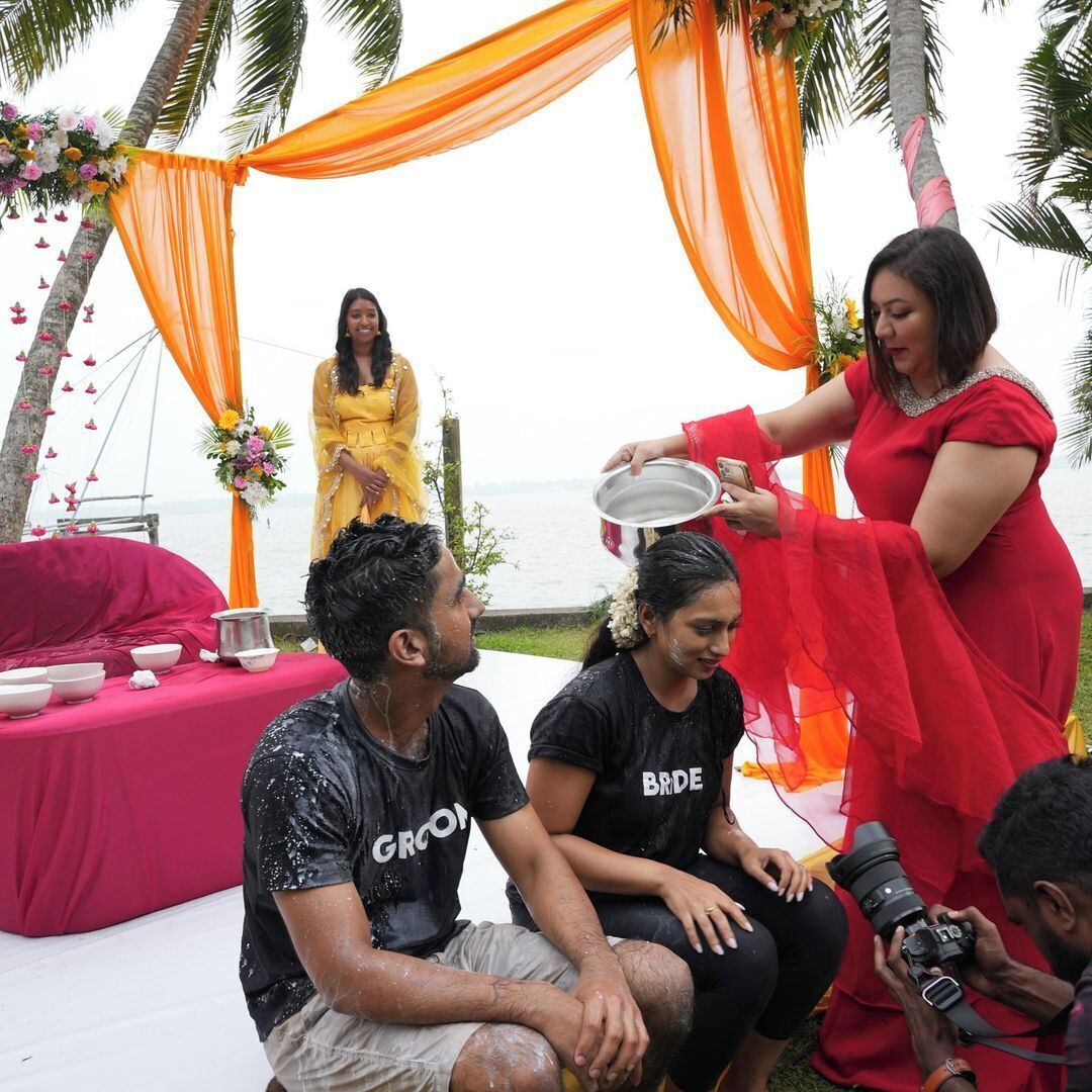 A wedding ceremony with a man and woman seated on a pink-covered table, surrounded by guests and decorations.