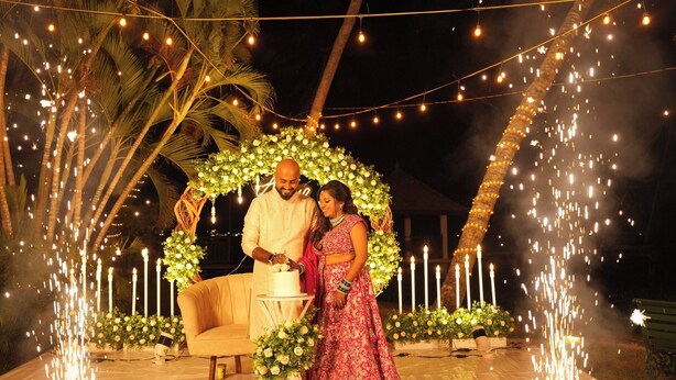 A couple standing under a floral arch with string lights, surrounded by candles and greenery. - Village Canopy | wedding venues kochi (Kochi)