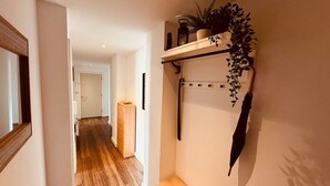 A hallway with wooden flooring, a bench, a wall-mounted shelf with plants, and a mirror.