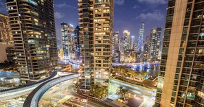 Paisagem urbana noturna com edifícios altos, uma ponte curva e um calçadão à beira-mar.