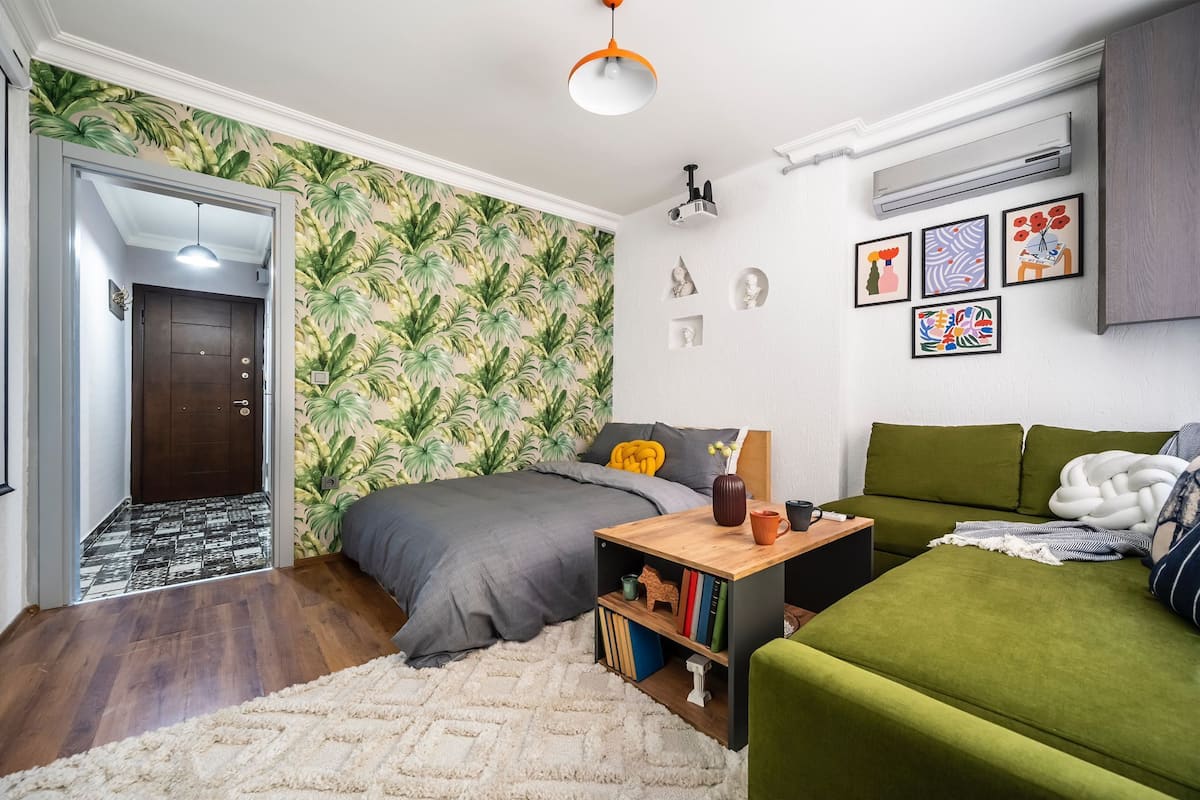 A compact bedroom with a bed, a green sofa, a small wooden side table, and a bookshelf.