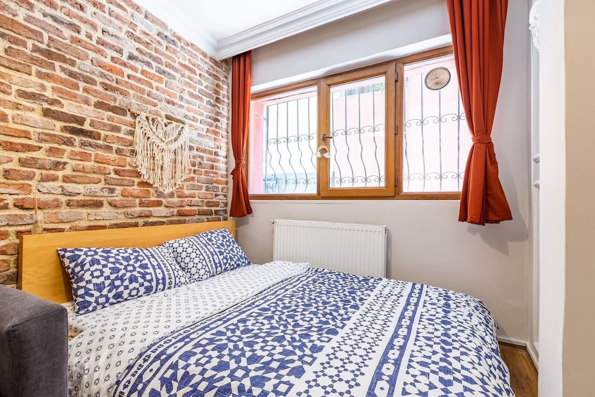 A bedroom with a brick wall, a wooden bed frame, patterned bedding, and a window with curtains.