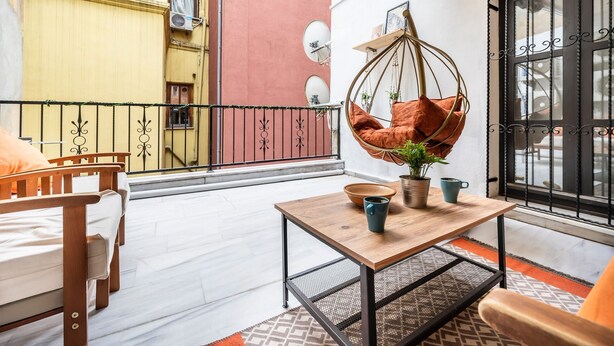 Un balcón con una hamaca colgante, una mesita con un cuenco y tazas, y vista a un edificio amarillo.