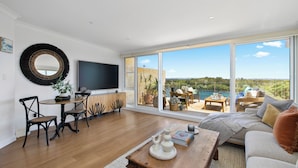 A spacious living area with a large window offering a view of the outdoors. There is a flat-screen TV mounted on the wall, a wooden coffee table with decorative items, and a comfortable seating area with a sofa and chairs. - Regatta 19 - Top Floor Luxury.  Spectacular Water Views | Breathtaking Sunsets  (Forster)