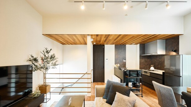 Ein modernes Wohnzimmer mit einem großen Flachbildfernseher, einer Couch und einem Couchtisch. Eine Treppe führt zu einer dunkleren Küchenzeile mit Kühlschrank und Mikrowelle.