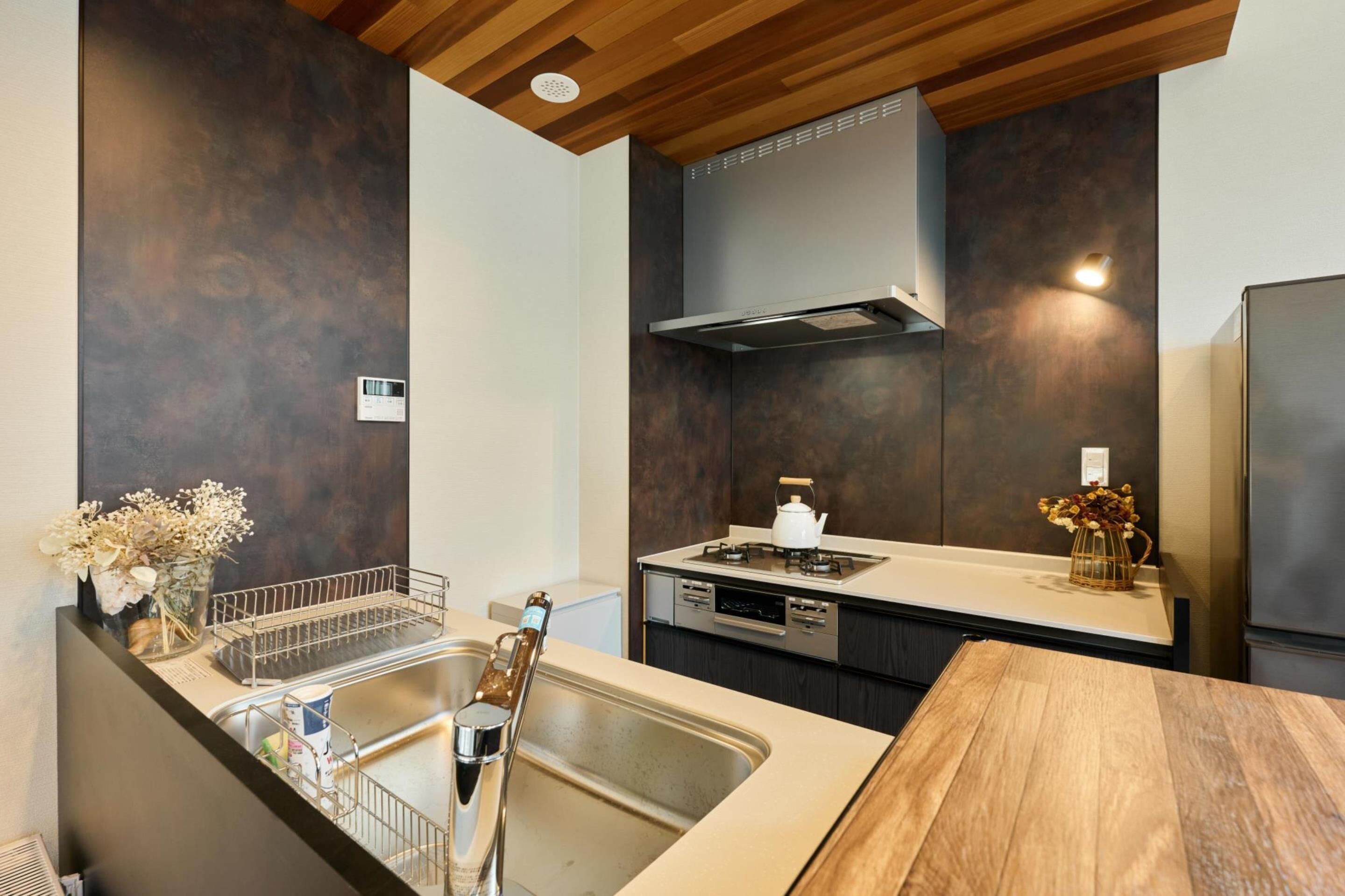 A modern kitchen with a wooden ceiling, dark wall panels, stainless steel appliances, and a wooden dining table.