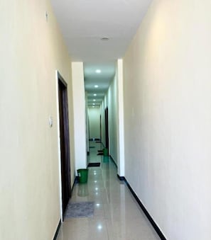 A long, narrow hallway with a shiny floor, white walls, and a green bucket on the left. - Hotel Shiv Aagya (Ujjain)