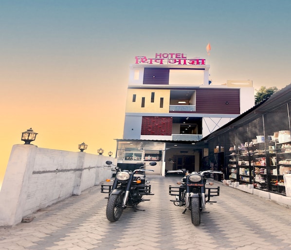 A hotel with a modern architectural design, featuring a prominent signboard and a paved entrance area with two motorcycles parked in front. - Hotel Shiv Aagya (Ujjain)