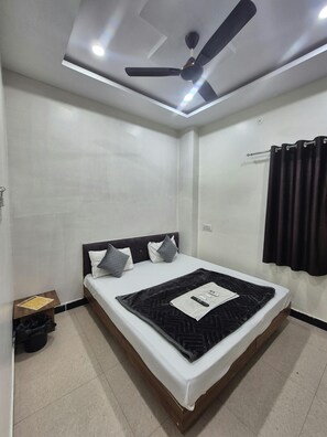 A neatly arranged bedroom with a bed, a ceiling fan, and a window with curtains. - Hotel Shiv Aagya (Ujjain)