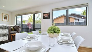 A dining area with a white table set for four, surrounded by white chairs. There is a potted plant on the table, and a window with a view of a house and trees. - Brand-new, townhouse - walking distance to Westfield Mall (Christchurch)
